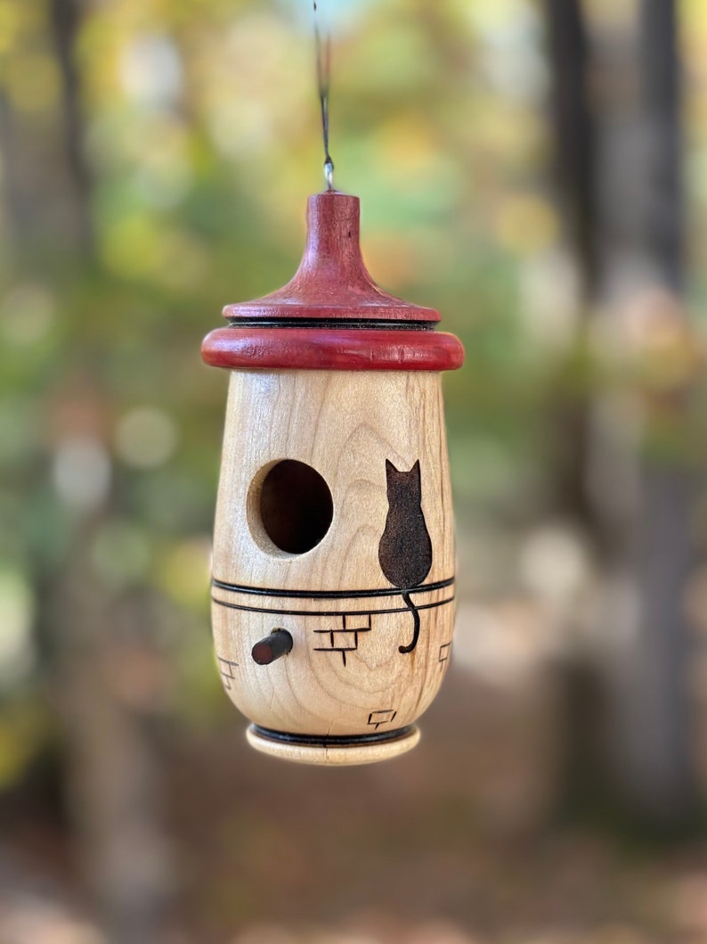 Puede incluir: Una casa para p&aacute;jaros de madera con un cuerpo de madera clara y una parte superior roja oscura. Una silueta de gato negro est&aacute; grabada en el lateral. La casa para p&aacute;jaros tiene un agujero de entrada redondo y una peque&ntilde;a percha. Cuelga de un hilo negro fino.