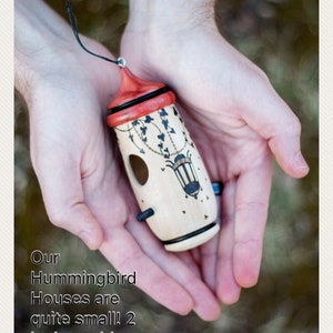 May include: A small, wooden hummingbird house with a red roof and decorative black accents. The house is approximately 2 inches wide and 5 inches tall. It features a small opening and a black hanging cord.
