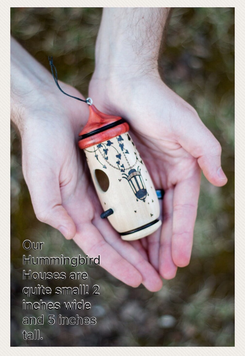 May include: A small, wooden hummingbird house with a red roof and decorative black accents. The house is approximately 2 inches wide and 5 inches tall. It features a small opening and a black hanging cord.