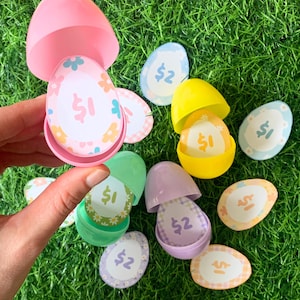 Puede incluir: Una colección de coloridos huevos de Pascua de plástico, algunos abiertos para revelar fichas en forma de huevo con diseños florales y cantidades en dólares. Los huevos son de varios colores, incluyendo rosa, amarillo, verde y morado, sobre un fondo de hierba verde.