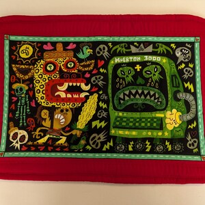 May include: Red quilted placemat with a green border featuring a colorful illustration of a monster with the text "Muerton 3000" and a creature with a leopard print.