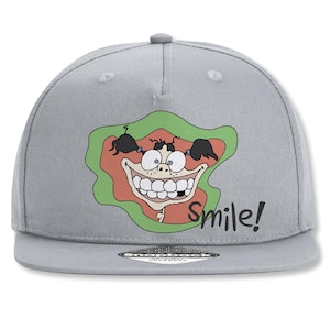 Könnte beinhalten: Graue Snapback-Kappe mit einem Cartoon-Gesicht, das ein großes, zahnreiches Grinsen, schwarze Zöpfe und blaue Augen zeigt. Das Gesicht befindet sich vor einem grünen und orangefarbenen Hintergrund, mit dem Wort "Smile!" in Schwarz.