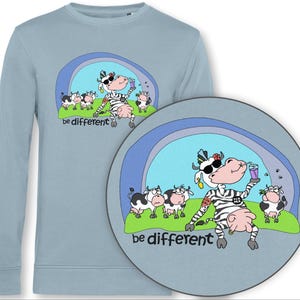 Könnte beinhalten: Hellblaues Langarm-Sweatshirt mit einem bunten Kuh-Cartoon-Design. Die Kuh trägt eine Sonnenbrille und Zebrastreifen und hält ein Getränk. Der Text "be different" steht unter dem Bild. Weitere Kühe sind im Hintergrund.