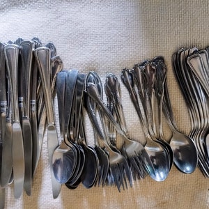 Puede incluir: Una colección de cubiertos de acero inoxidable, que incluye cuchillos, cucharas y tenedores, dispuestos sobre un paño de color claro. Los cubiertos tienen una superficie brillante y reflectante, y los mangos presentan varios diseños decorativos.