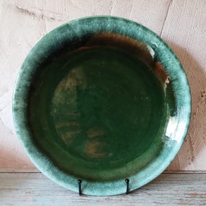 Può includere: Un piatto rotondo in ceramica con un centro verde scuro lucido e un bordo verde più chiaro. Il piatto è esposto su un supporto in metallo nero, che mette in risalto il suo design e le variazioni di colore. La superficie del piatto riflette la luce.