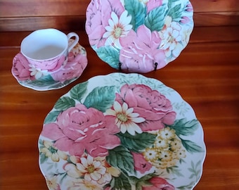 May include: A floral-themed tea set featuring a teacup, saucer, and two plates. The set showcases a pink rose and floral design with green leaves on a white background. The plates have a scalloped edge.