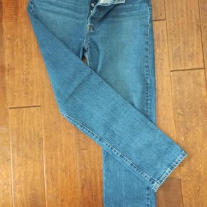 May include: Blue denim jeans with a button fly, pockets, and a classic straight leg. The jeans are laid flat on a wooden surface, showcasing their design and construction. The denim has a medium wash.