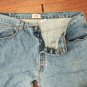 Könnte beinhalten: Hellblaue Jeans aus Denim mit Knopfleiste und klassischem Fünf-Taschen-Design. Die Jeans sind leicht verblichen und weisen Gebrauchsspuren auf. Das Etikett lautet "501".