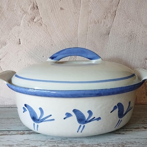 Puede incluir: Una fuente de cerámica blanca con tapa, con detalles azules e ilustraciones de pájaros. La fuente tiene un asa azul y un borde azul. Es un artículo de menaje de cocina.