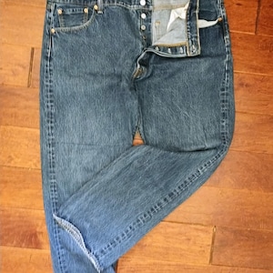 May include: Blue denim jeans with a button fly, visible pockets, and a classic straight-leg fit. The jeans are laid flat on a wooden surface, showcasing their details and construction. The denim has a faded appearance.