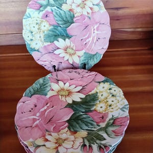 May include: A set of floral-patterned plates featuring pink roses, white daisies, and yellow flowers on a white background. The plates have a scalloped edge and are arranged on a wooden surface. The plates are a mix of pink, white, yellow, and green.