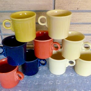 May include: A selection of ceramic mugs in shades of yellow, cream, blue, and red. Each mug features a rounded handle and a simple, classic design. The mugs are stacked together, displaying their different colours.