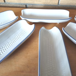 May include: White ceramic serving dishes with a blue trim. The dishes are shaped like corn on the cob, with textured patterns on the inside. The dishes are arranged on a wooden surface.