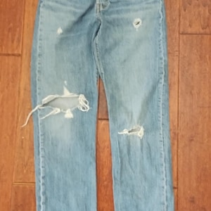 May include: Light blue denim jeans with distressed details, including rips and faded areas. The jeans have a button fly and classic five-pocket design. The garment is displayed on a wooden surface.