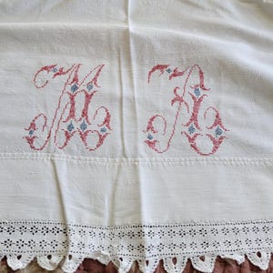 May include: White linen sheet with embroidered initials "A B" in red and blue. The sheet features a delicate lace trim along the bottom edge. The fabric is slightly wrinkled, suggesting it is vintage.