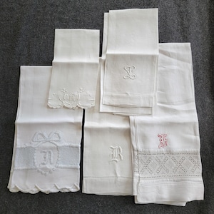 Può includere: Una collezione di asciugamani di lino bianco, alcuni piegati e altri spiegati, con dettagli ricamati. Gli asciugamani presentano vari disegni, tra cui motivi floreali, monogrammi e bordi decorativi. Gli asciugamani sono disposti su una superficie grigia.