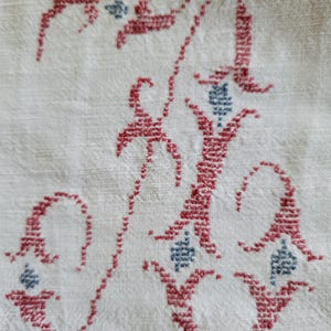 May include: Close-up of a white fabric with a detailed embroidered letter "A". The letter is in a script style, stitched in red thread with small blue accents. The fabric has a textured appearance.
