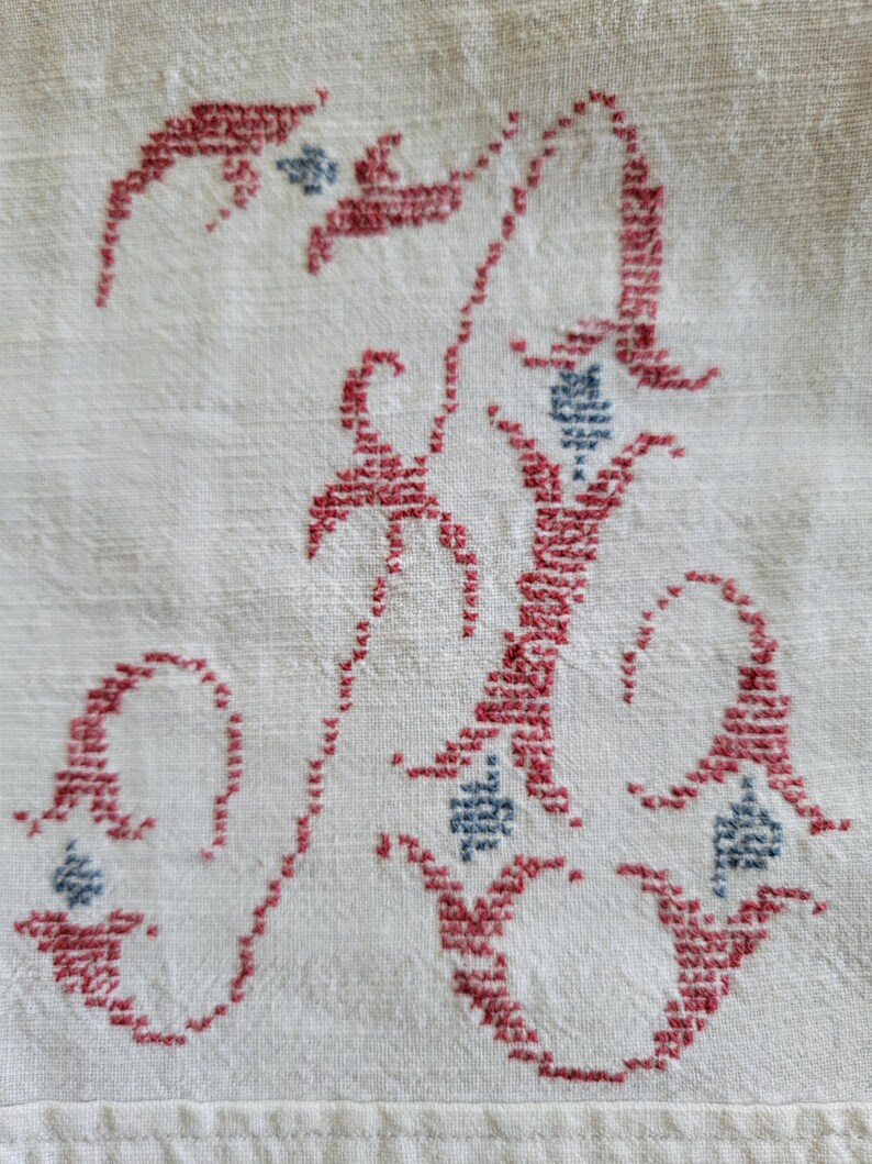 May include: Close-up of a white fabric with a detailed embroidered letter "A". The letter is in a script style, stitched in red thread with small blue accents. The fabric has a textured appearance.