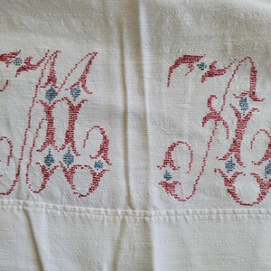 May include: Cream-colored fabric with two embroidered letters, possibly initials. The letters are a deep red with blue diamond-shaped accents. The embroidery is detailed and appears to be vintage or handmade, suggesting a textile item.