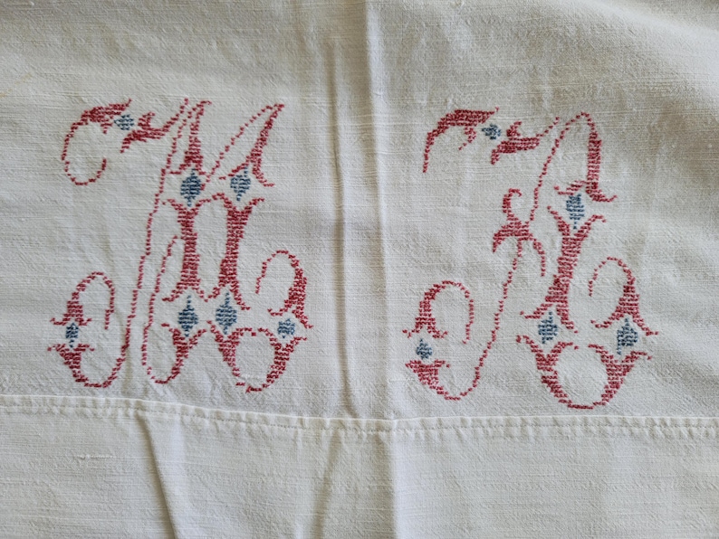 May include: Cream-colored fabric with two embroidered letters, possibly initials. The letters are a deep red with blue diamond-shaped accents. The embroidery is detailed and appears to be vintage or handmade, suggesting a textile item.