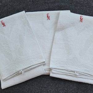 以下が含まれることがあります： 織り目模様と赤い刺繍のイニシャルが入った、3つの折り畳まれた白いリネンナプキン。ナプキンは濃い灰色の表面に積み重ねられています。イニシャルは様式化されたスクリプトです。