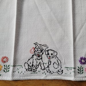 Könnte beinhalten: Weißes Küchentuch mit gestickten Welpen und Blumen. Zwei schwarz-weiße Welpen sind in der Mitte gestickt, mit bunten Blumen auf jeder Seite. Das Handtuch hat ein strukturiertes Gewebe.