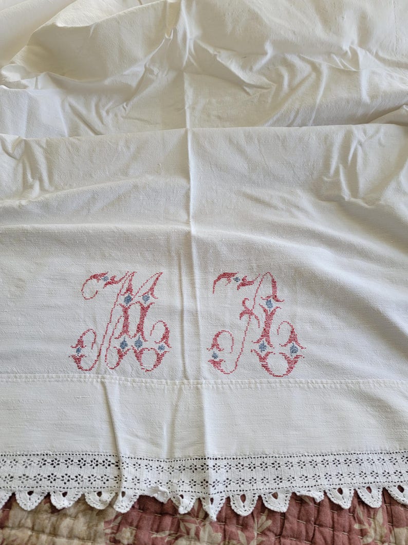 May include: White linen sheet with embroidered initials "A B" in red and blue. The sheet features a delicate lace trim along the bottom edge. The fabric is slightly wrinkled, suggesting it is vintage.