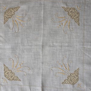 May include: A light gray rectangular textile featuring a repeating pattern of embroidered floral motifs in beige thread. The design includes intricate openwork lace-like corners and delicate curved stems with small dots, finished with a hemstitched border.