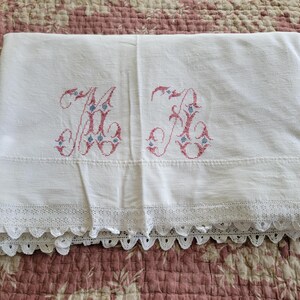 May include: White linen sheet with embroidered pink and blue initials. The sheet features a decorative lace trim along the bottom edge. The sheet is folded and displayed on a floral patterned surface.