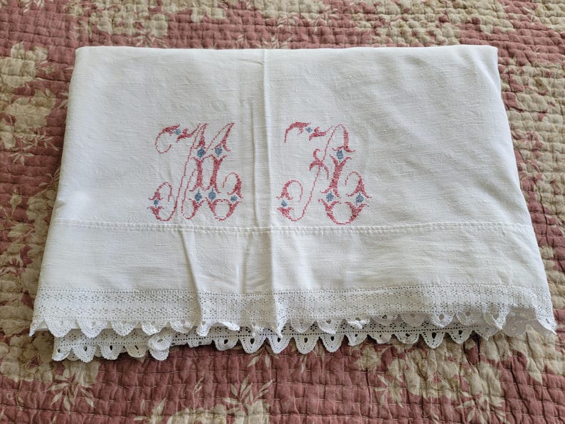 May include: White linen sheet with embroidered pink and blue initials. The sheet features a decorative lace trim along the bottom edge. The sheet is folded and displayed on a floral patterned surface.