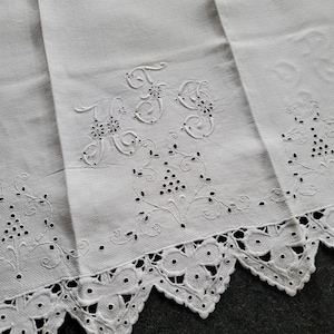 Puede incluir: Paño de lino blanco con iniciales bordadas "FS" y diseños florales. Los bordes presentan un borde festoneado con detalles recortados. El bordado incluye pequeños puntos negros, que añaden contraste a la tela blanca. Un textil vintage.