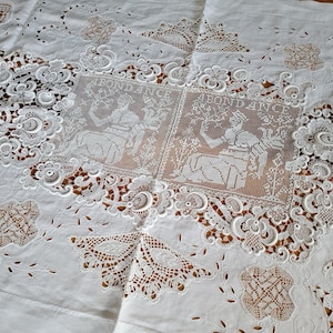 Op de afbeelding: Witte vintage tafelkleed met ingewikkelde kanten details. Voorzien van ajourwerk en geborduurde bloemen- en geometrische patronen. Bevat twee rechthoekige panelen met het woord "ABONDANCE" en een zittende figuur.