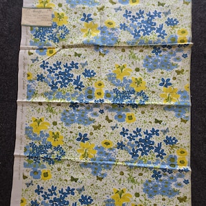 Può includere: Un pezzo di tessuto piegato con un motivo floreale. Il design presenta fiori blu e gialli, foglie verdi e farfalle su sfondo bianco. Il tessuto ha un'etichetta attaccata.