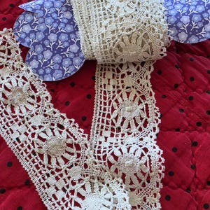 May include: A roll of off-white crochet lace with a floral design, paired with a blue floral paper cutout, rests on a red quilted surface with black polka dots. The lace is intricately detailed and appears to be vintage.