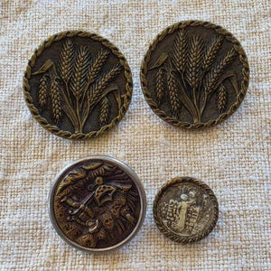May include: Four antique buttons with detailed designs. Two round buttons feature wheat stalks with a rope-like border. Another button has a detailed relief design, and the smallest button has a relief design with two figures.
