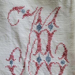 May include: Close-up of a white linen textile with a cross-stitched letter "M". The embroidery is in red and blue, featuring a decorative design with diamond-shaped accents. The fabric has a subtle texture.
