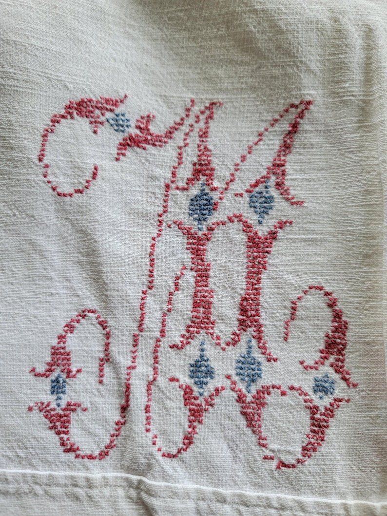 May include: Close-up of a white linen textile with a cross-stitched letter "M". The embroidery is in red and blue, featuring a decorative design with diamond-shaped accents. The fabric has a subtle texture.