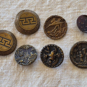 May include: A collection of antique brass buttons with intricate designs. The buttons feature various motifs, including geometric patterns, birds, floral elements, and a moon with a face. The buttons are arranged on a textured, beige fabric.