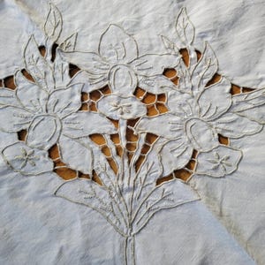 Pode incluir: Close-up de um bordado floral branco em tecido claro. O bordado apresenta um buquê de flores detalhado com seções recortadas revelando um fundo marrom. O tecido tem uma aparência ligeiramente texturizada.