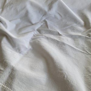 May include: Close-up of a white, textured fabric with soft folds and subtle shadows. The fabric appears to be a sheet or similar textile, with visible seams along the bottom edge. The overall impression is of a clean, neutral surface.