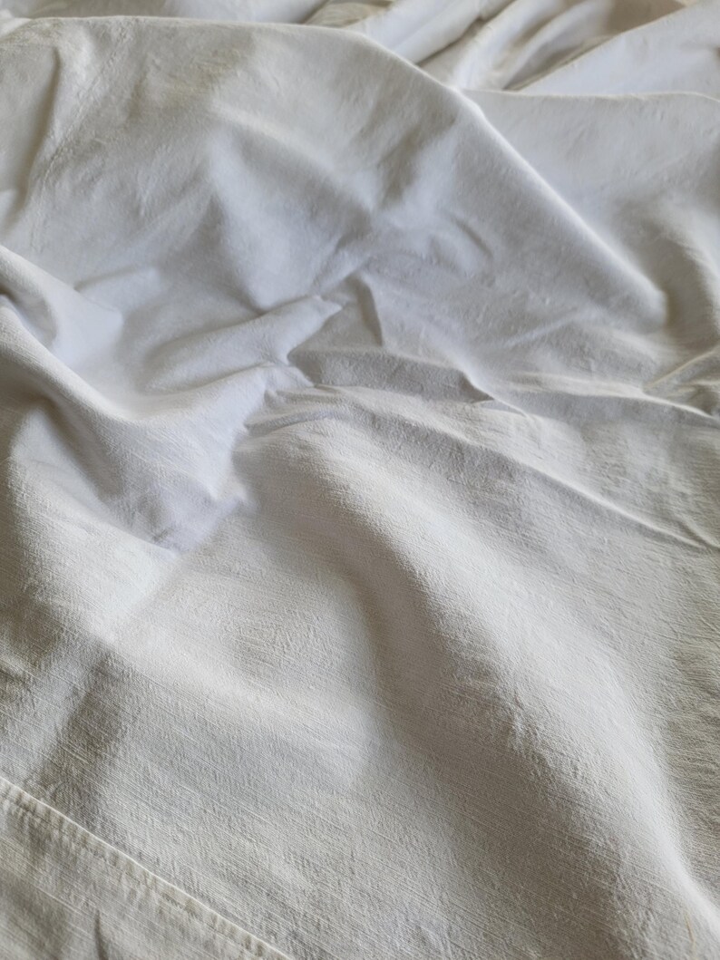 May include: Close-up of a white, textured fabric with soft folds and subtle shadows. The fabric appears to be a sheet or similar textile, with visible seams along the bottom edge. The overall impression is of a clean, neutral surface.