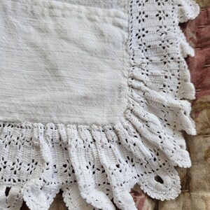 May include: Close-up of a white linen apron with a crocheted scalloped edge. The apron has a pocket and a textured surface. The crocheted trim adds a delicate, handmade touch to the apron's design.