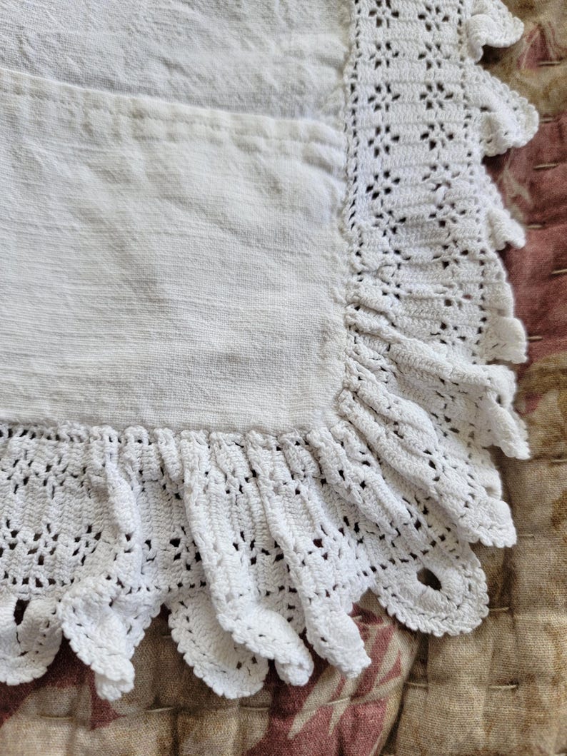 May include: Close-up of a white linen apron with a crocheted scalloped edge. The apron has a pocket and a textured surface. The crocheted trim adds a delicate, handmade touch to the apron's design.