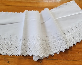 May include: White eyelet fabric with a scalloped edge. The fabric is folded, revealing the intricate floral cut-out design along the border. The eyelet trim adds a delicate touch to this textile.
