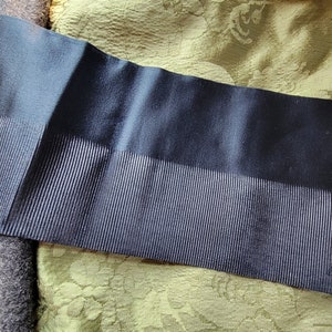May include: A black fabric trim with a ribbed texture. The trim is approximately 2.5 cm wide and has a smooth, satin-like finish.