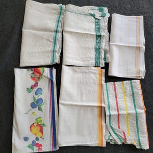 Puede incluir: Se muestran seis paños de cocina blancos. Algunos tienen rayas verdes, naranjas, amarillas y rojas. Un paño presenta un diseño de frutas con melocotones, ciruelas y bayas. Los paños están doblados y colocados sobre una superficie oscura.