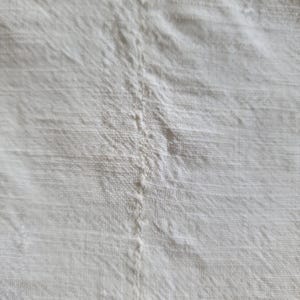 May include: Close-up of a white linen fabric with visible texture and a central seam. The fabric appears to be slightly wrinkled, with subtle variations in tone, suggesting a natural, unbleached material. The overall impression is of a soft, natural textile.