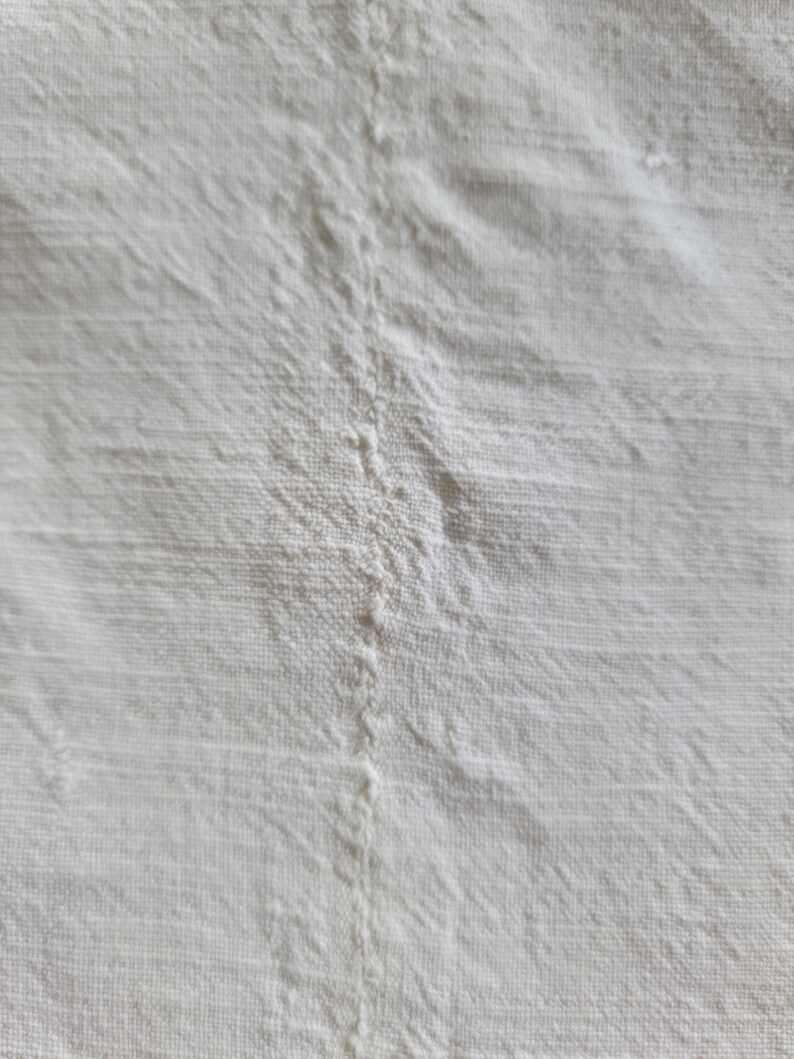 May include: Close-up of a white linen fabric with visible texture and a central seam. The fabric appears to be slightly wrinkled, with subtle variations in tone, suggesting a natural, unbleached material. The overall impression is of a soft, natural textile.