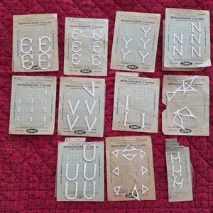 May include: A collection of vintage DMC monogram embroidery patterns. The patterns feature white plastic letters and shapes on aged paper cards. The cards are arranged on a red quilted surface.