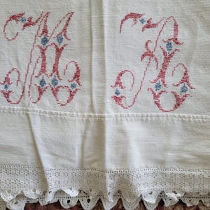 May include: Cream-colored linen textile with embroidered red and blue initials "M" and "K". The initials are embellished with floral details. The bottom edge features a delicate white lace trim. This vintage textile is suitable for home decor.